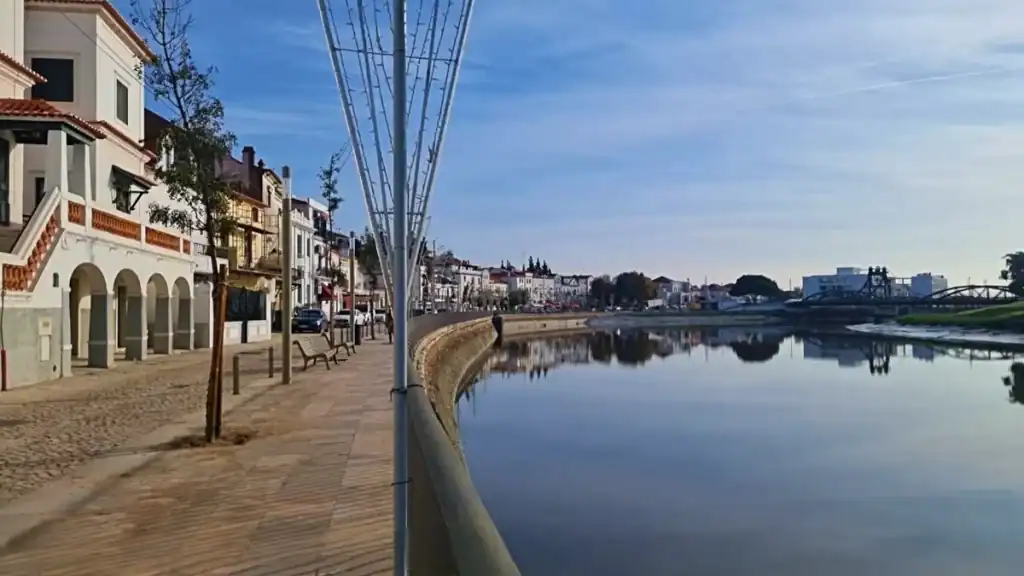 Calm river reflecting whitewashed buildings along a wide pedestrian promenade with benches and trees