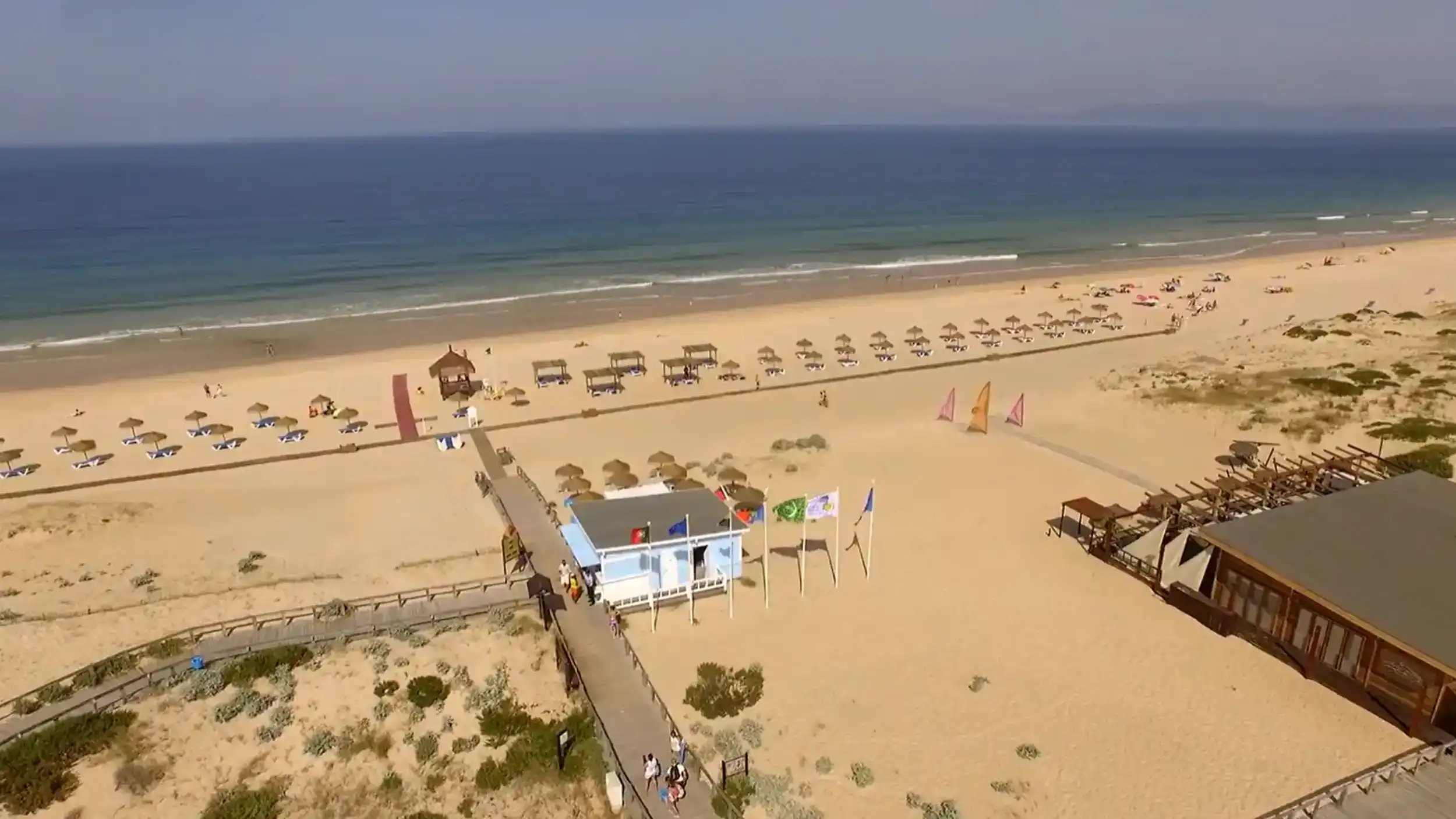 Aerial view of a wide sandy beach with thatched umbrellas, a boardwalk, and the Atlantic beyond
