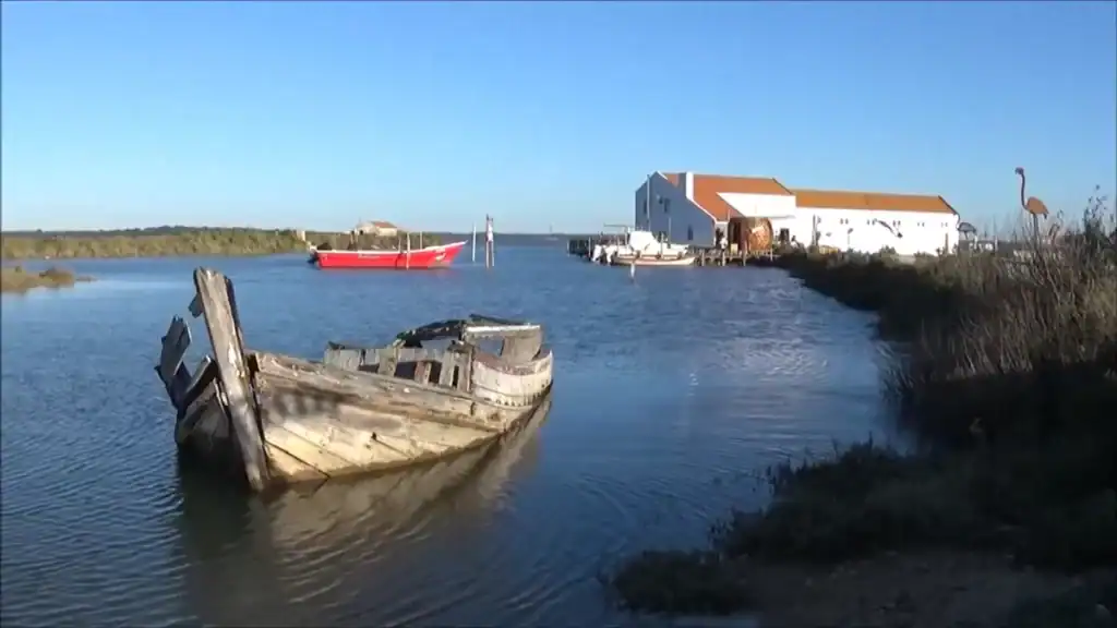 Decaying wooden boat hull half-submerged in a calm estuary channel beside a whitewashed warehouse
