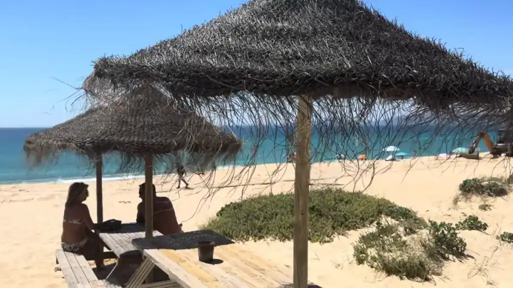 Couple sitting under a thatched straw parasol on golden sand with turquoise Atlantic water behind