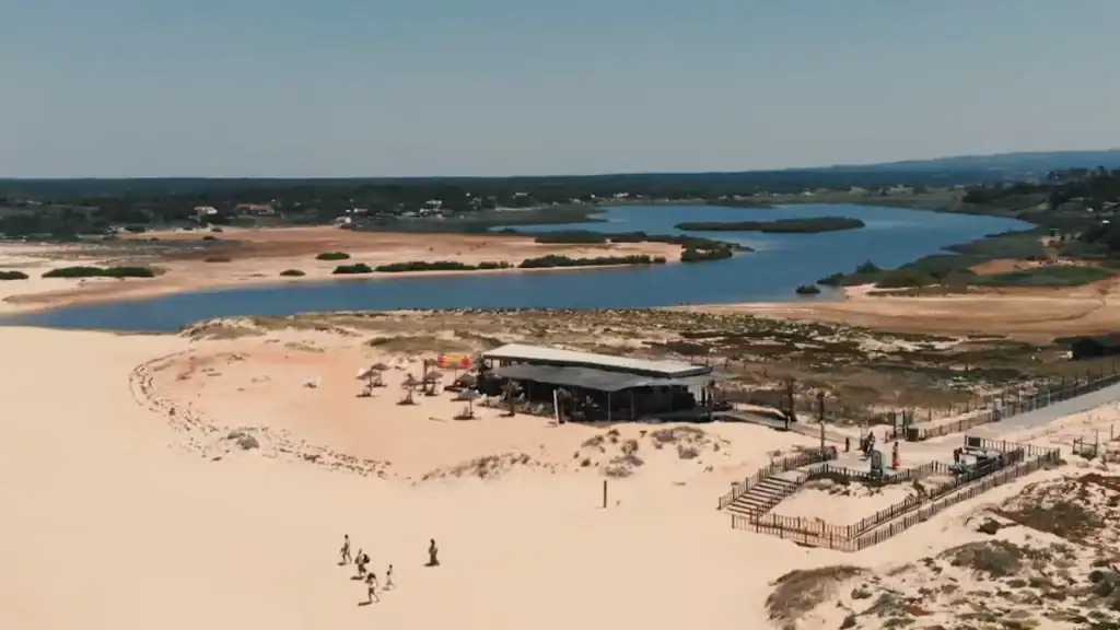 A beach bar with straw parasols on sand dunes beside a lagoon with pine-covered banks