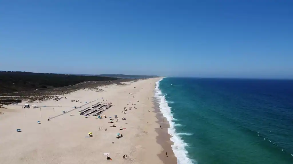 Wide sandy Melides beach with sunbathers, parasols, and Atlantic surf stretching to the horizon