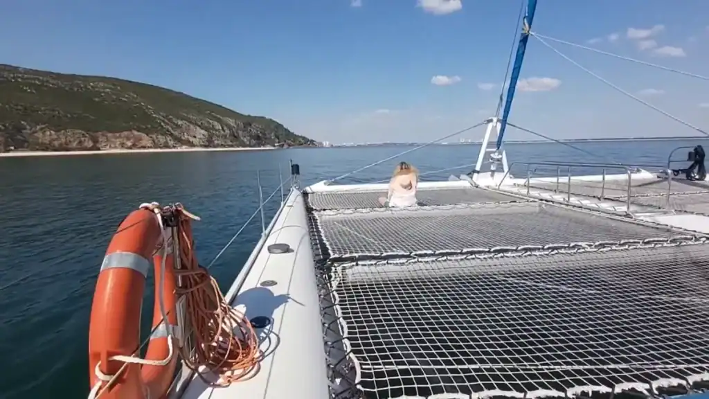 Woman sitting on catamaran netting as the boat passes rocky coastal cliffs
