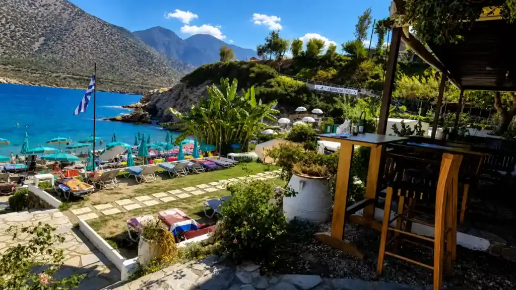 Beach bar terrace overlooking Karavostasi cove with Greek flag and mountain backdrop