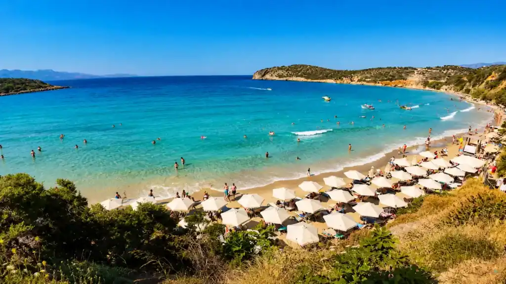 Swimmers and white umbrellas filling Voulisma Bay, a busy summer day at the beaches near Agios Nikolaos