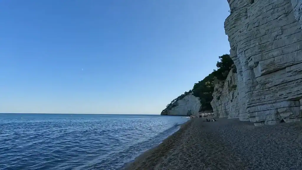 Towering layered cliffs casting shadow over a pebble shore at Vignanotica beach.
