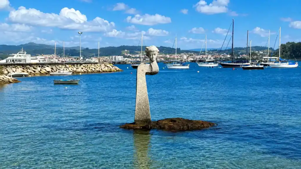 Stone figure sculpture rising from the water in O Grove harbour with yachts moored behind