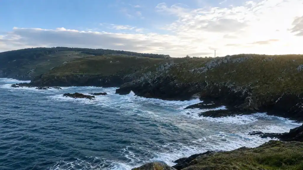Waves breaking against jagged rocks below green headlands on the Galician coast