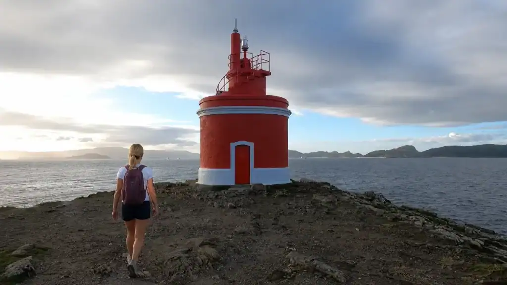 Woman approaching the red Faro de Punta Robaleira lighthouse on a rocky headland at dusk