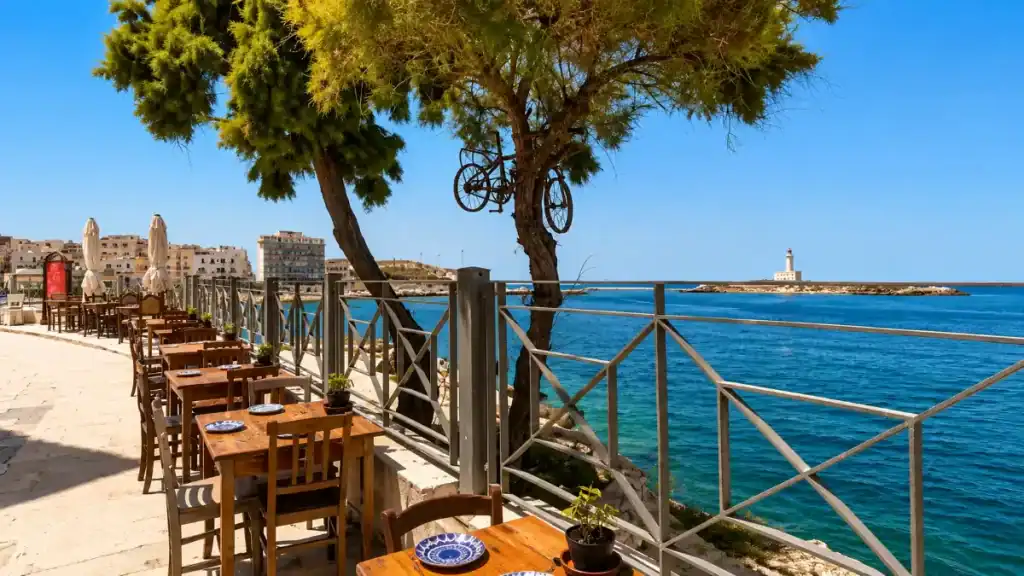 Wooden tables line a seafront terrace in Vieste with the lighthouse on Sant'Eufemia island.