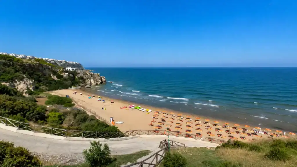 Orange beach umbrellas lined across the sandy crescent of Spiaggia di Peschici beneath the whitewashed clifftop town.
