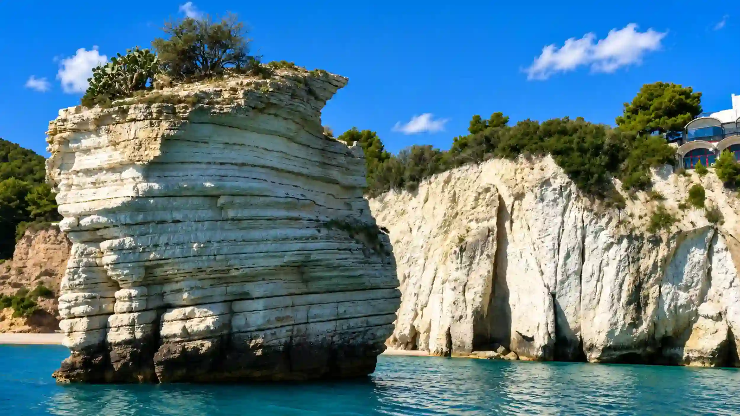 Towering limestone sea stack rising from turquoise water near Vieste, comparing East vs North Gargano coastlines.