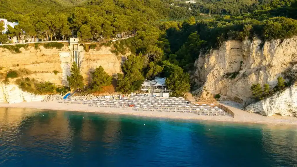 White umbrellas lined along the pebble shore beneath limestone cliffs at Baia delle Zagare.