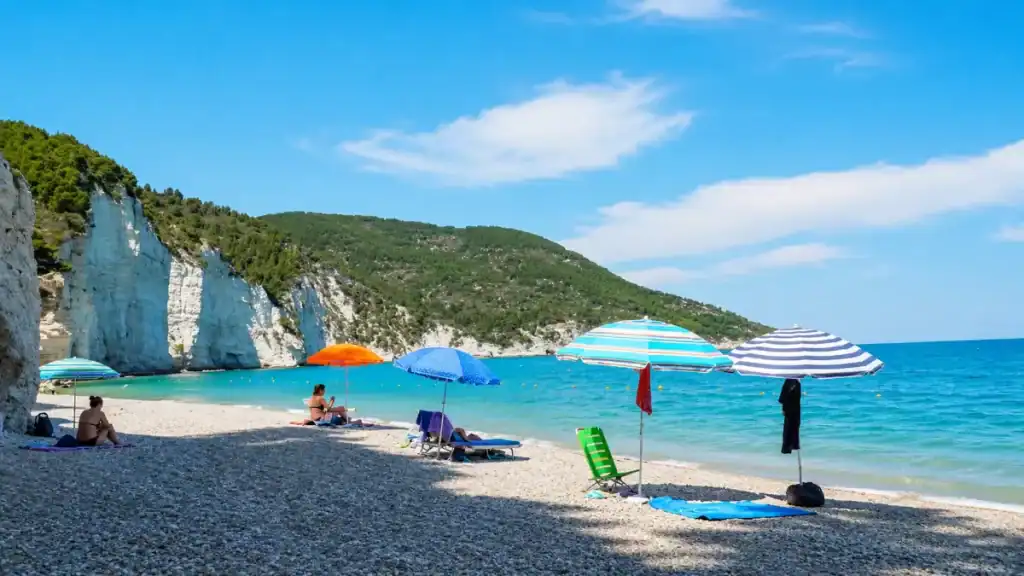 Striped parasols on pebbled shore below white cliffs at Vignanotica, a standout Gargano Puglia beach.