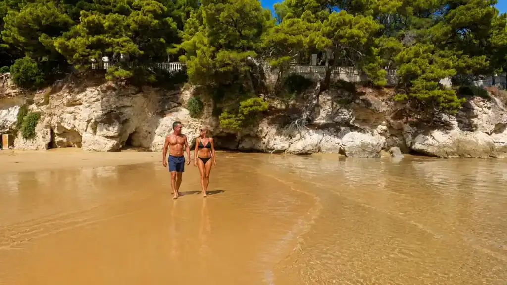 Couple walking along the wet sand at Baia San Felice beneath low pine-topped cliffs.
