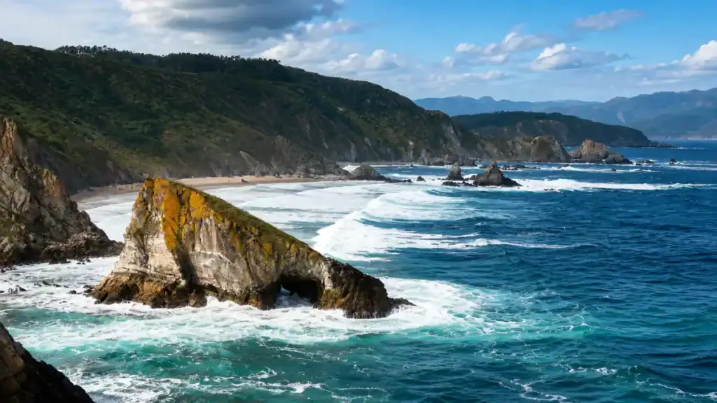Lichen-covered arched sea rock rising from crashing Atlantic waves along the wild Galician coastline with forested cliffs beyond