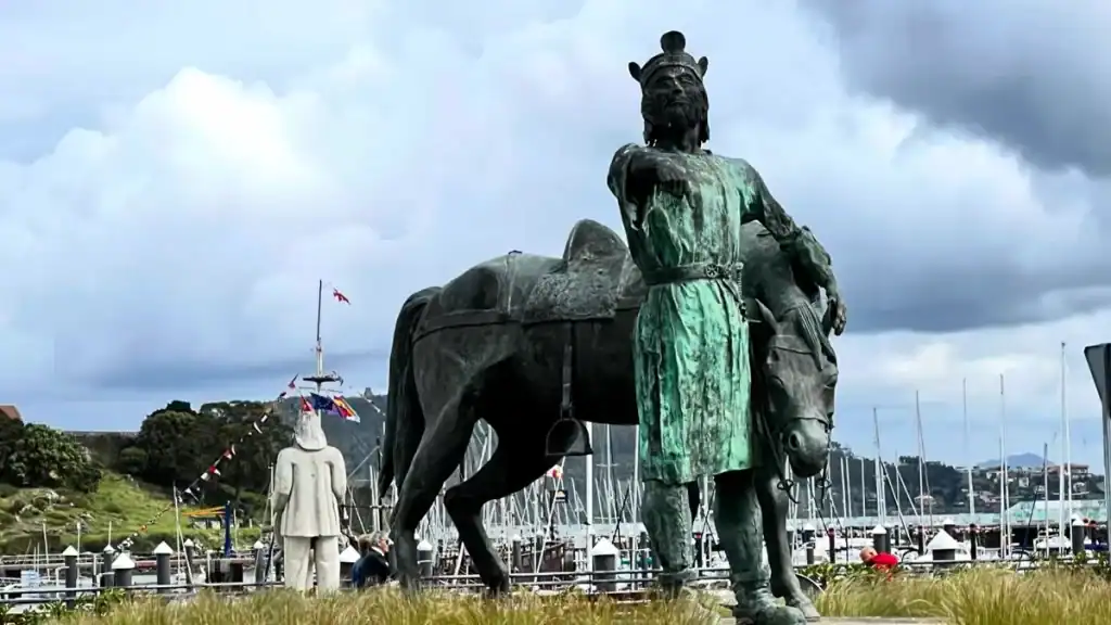 Bronze statue of King Alfonso IX standing beside a horse on Baiona's waterfront with the marina and hillside fortress behind