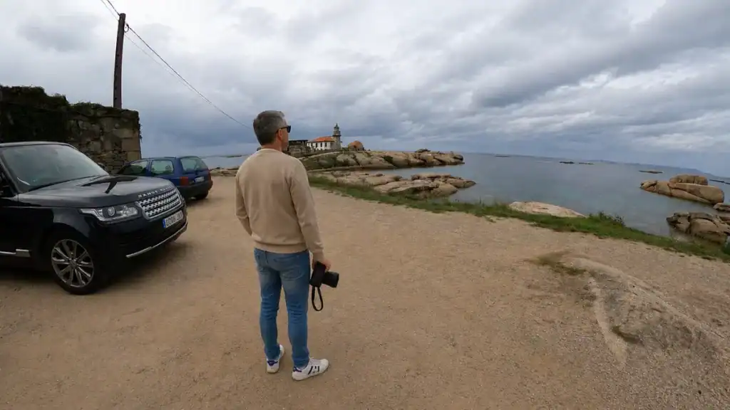 Photographer standing on a rocky headland near Baiona looking toward a small white lighthouse on granite outcrops by the sea