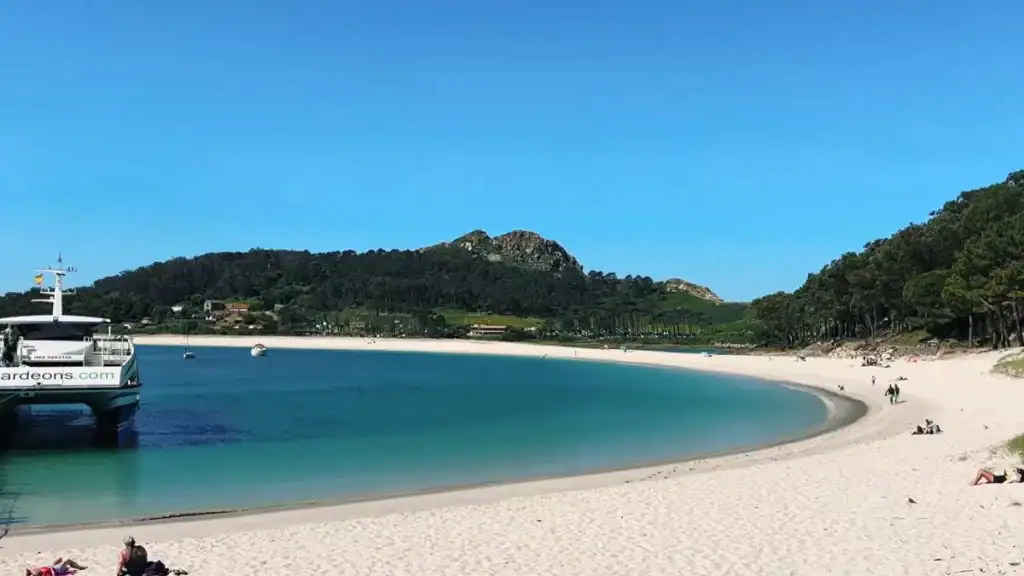 Curved white sand shoreline of Praia de Rodas on the Cíes Islands with turquoise lagoon water and a ferry docked at the pier