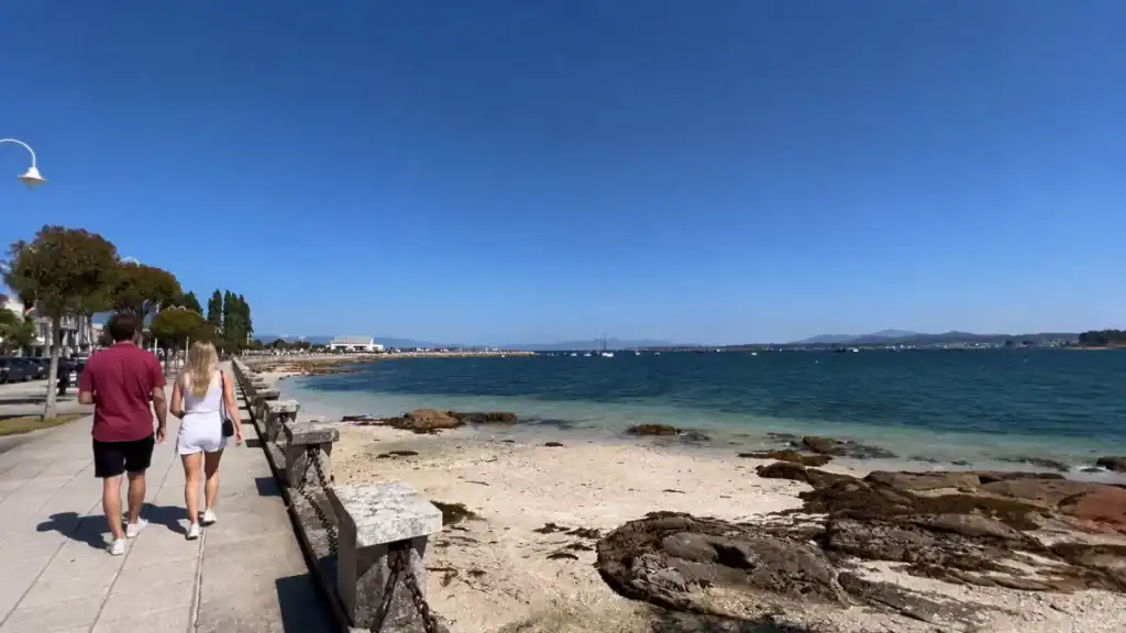 Couple walking the seafront path beside rocky tidal shore and turquoise waters at O Grove