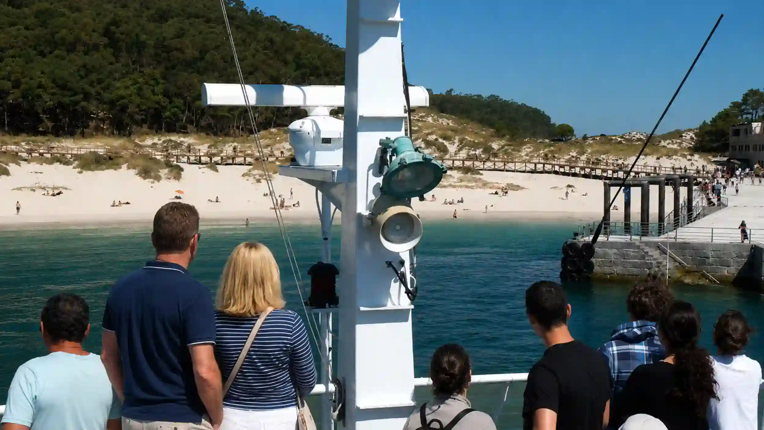 Passengers on ferry deck approaching Cíes Islands pier with white sand beach beyond
