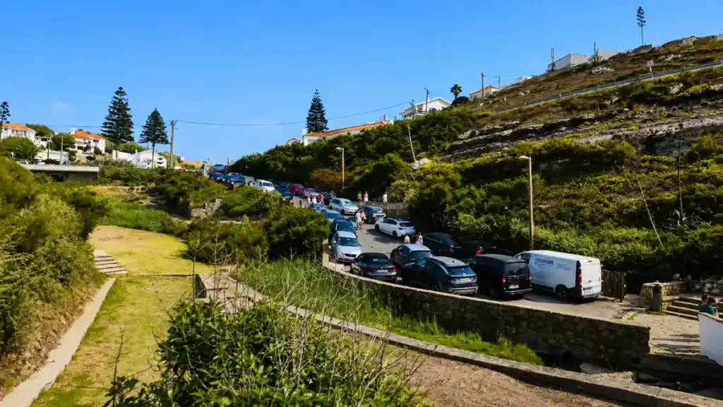 Packed roadside car park on a hillside road leading down to a small coastal village on a sunny day