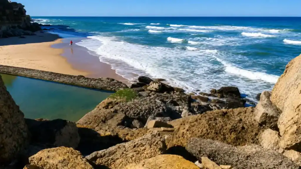 Natural seawater pool at Azenhas do Mar beside a sandy beach with a lone walker at the shoreline