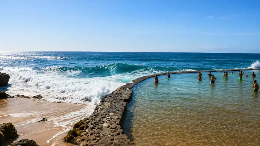 Swimmers in a clear tidal pool separated from crashing Atlantic waves by a low curved stone wall