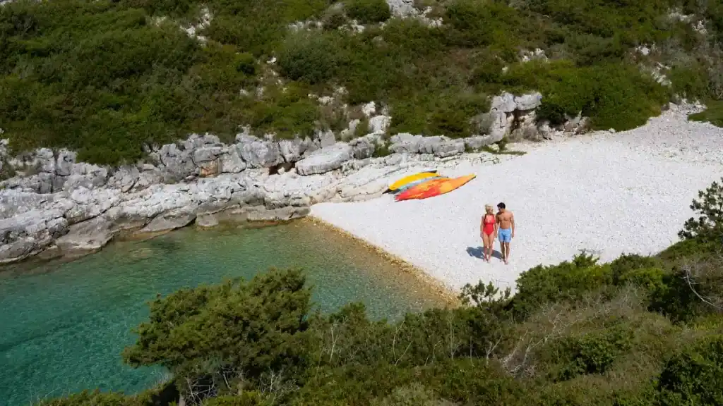 Kayaks rest on the white pebbles of secluded Avlaki Beach as a couple strolls by.