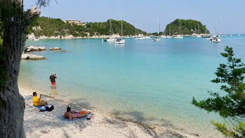 Sunbathers rest on a shaded pebble shore at Kanoni, boats scattered across the bay.