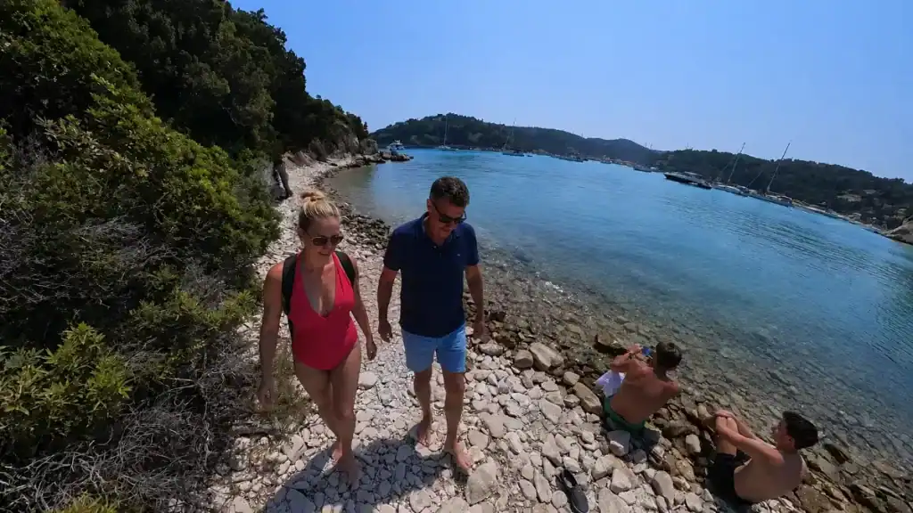 Two walkers cross the rocky shoreline path near Komeno with Gaios harbour beyond.
