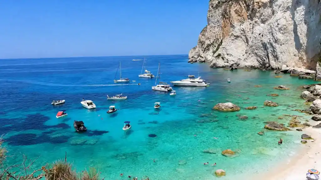 Boats anchored beneath tall white cliffs above the turquoise cove at Erimitis