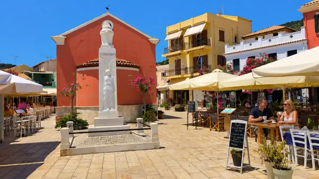 A white memorial statue standing before a terracotta church in Gaios main square, surrounded by cafรฉ tables