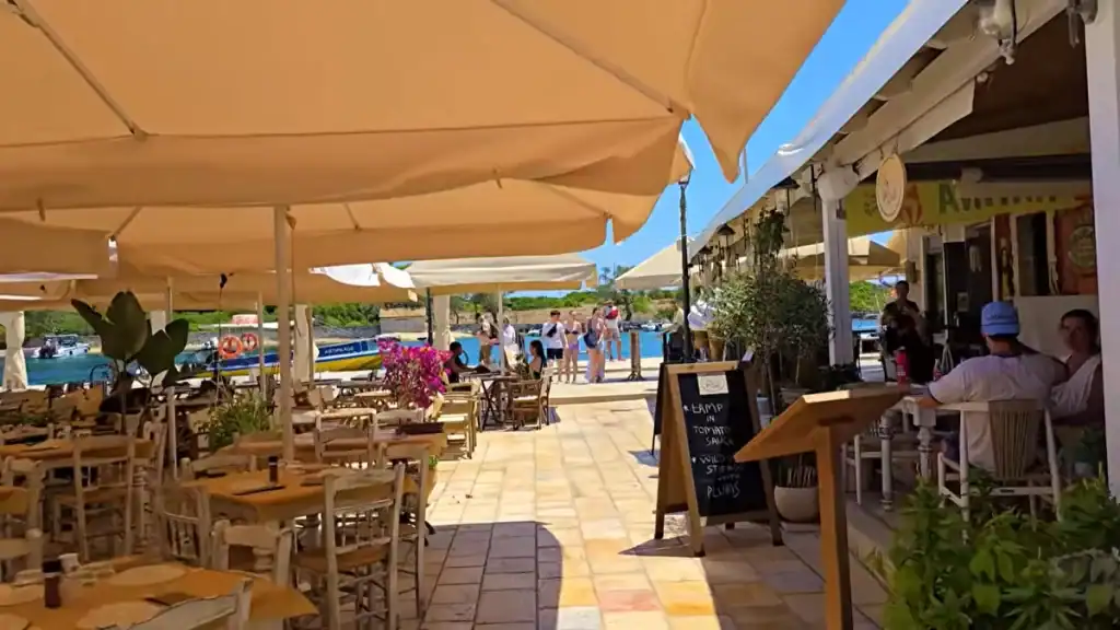 Diners seated under canvas umbrellas at a waterfront taverna with a chalkboard menu and boats moored beyond