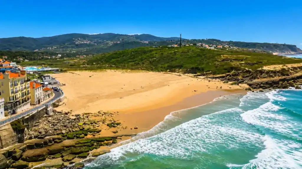 Wide sandy beach at Praia das Maçãs with mossy rocks and colourful seafront buildings below green hills