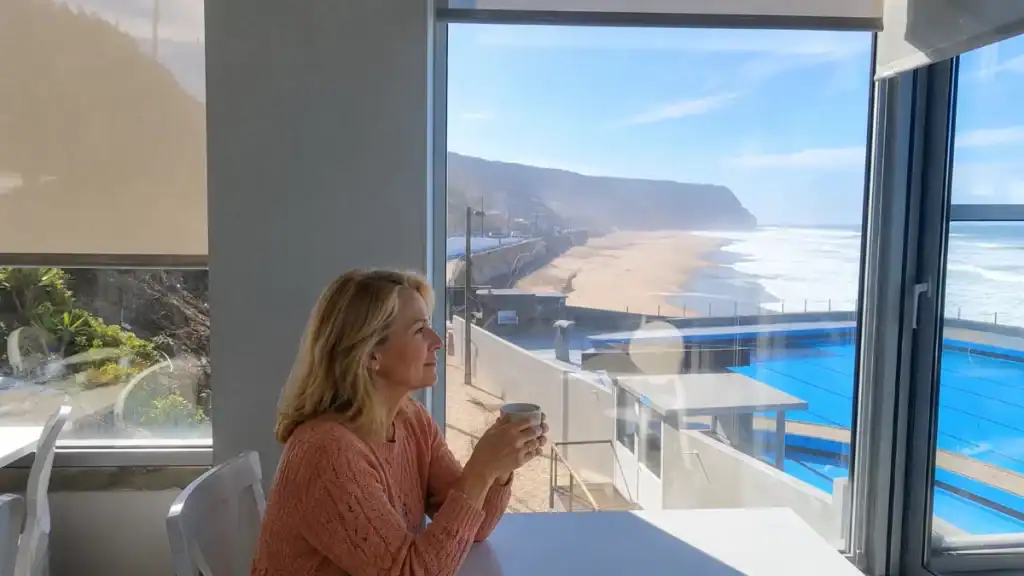 Woman holding a coffee cup gazing through large windows at Praia das Maçãs beach and cliffs, Colares
