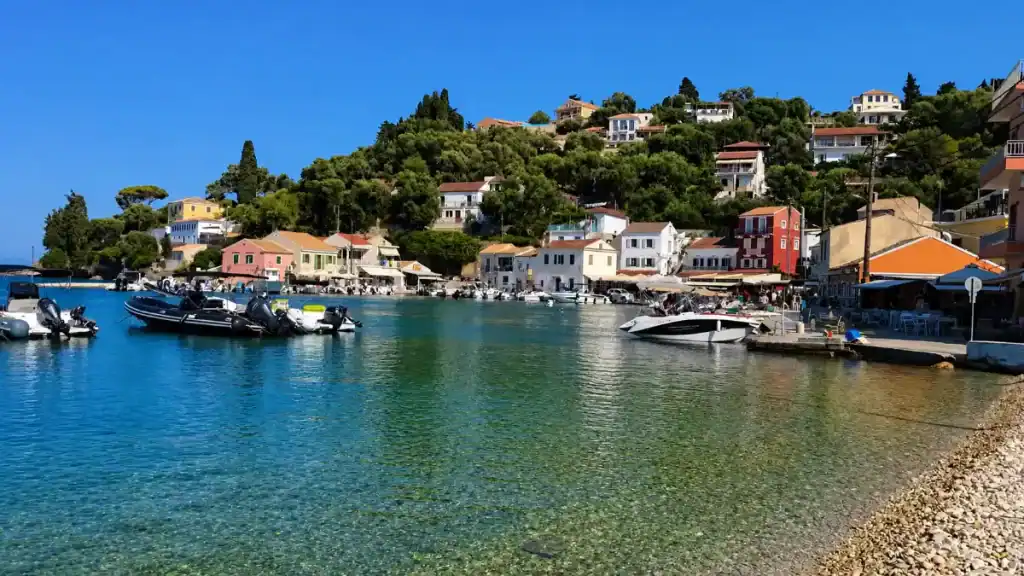 Small boats float in the clear turquoise waters of Loggos harbour beneath a pine-covered hillside.