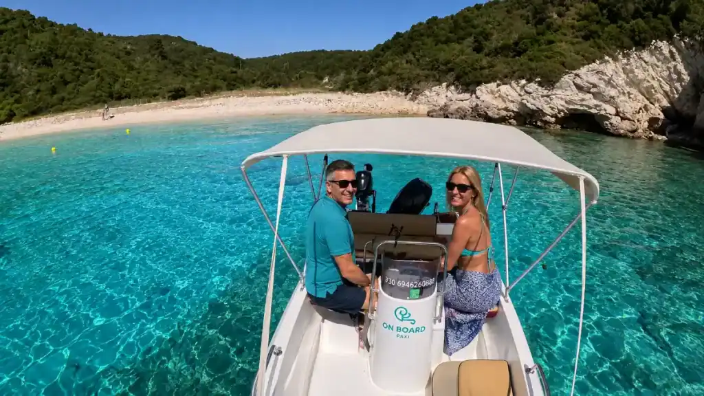 Couple piloting a rental boat toward a secluded pebble cove, is Antipaxos worth visiting for its hidden coves.