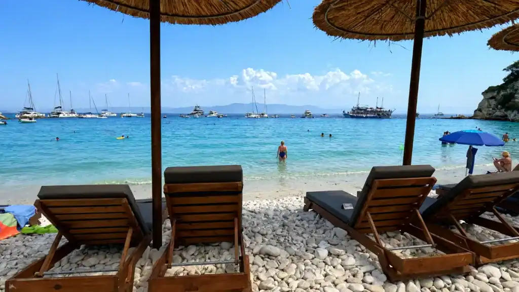 Wooden sunbeds on pebbled shore face anchored yachts scattered across Voutoumi Bay.