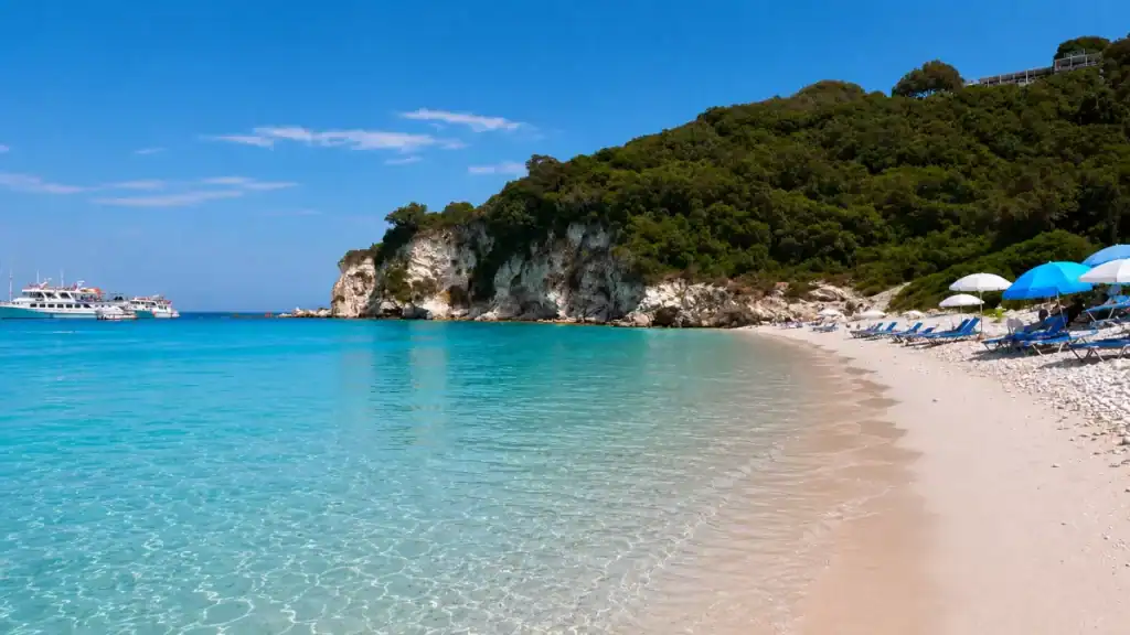 Empty sunbeds and umbrellas line the pale shore of Voutoumi Beach beneath forested cliffs.