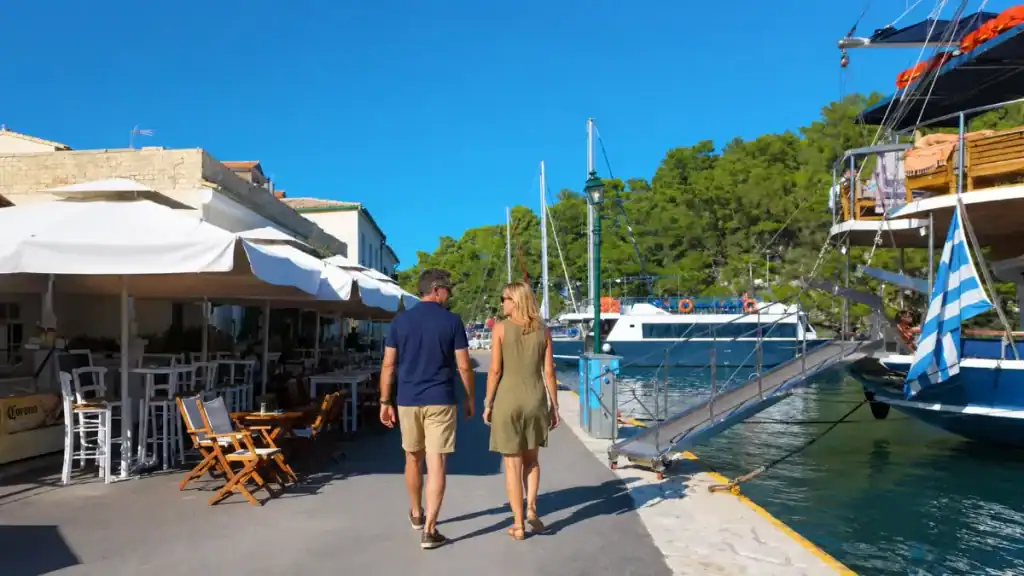 A couple walks the promenade past cafe umbrellas and yachts in Gaios harbour