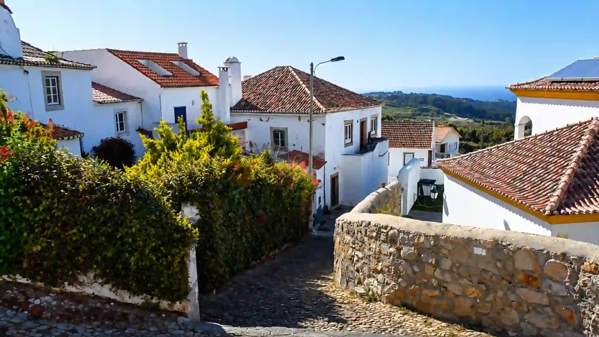Cobbled street winds past whitewashed houses in Penedo, Sintra, toward distant hills.