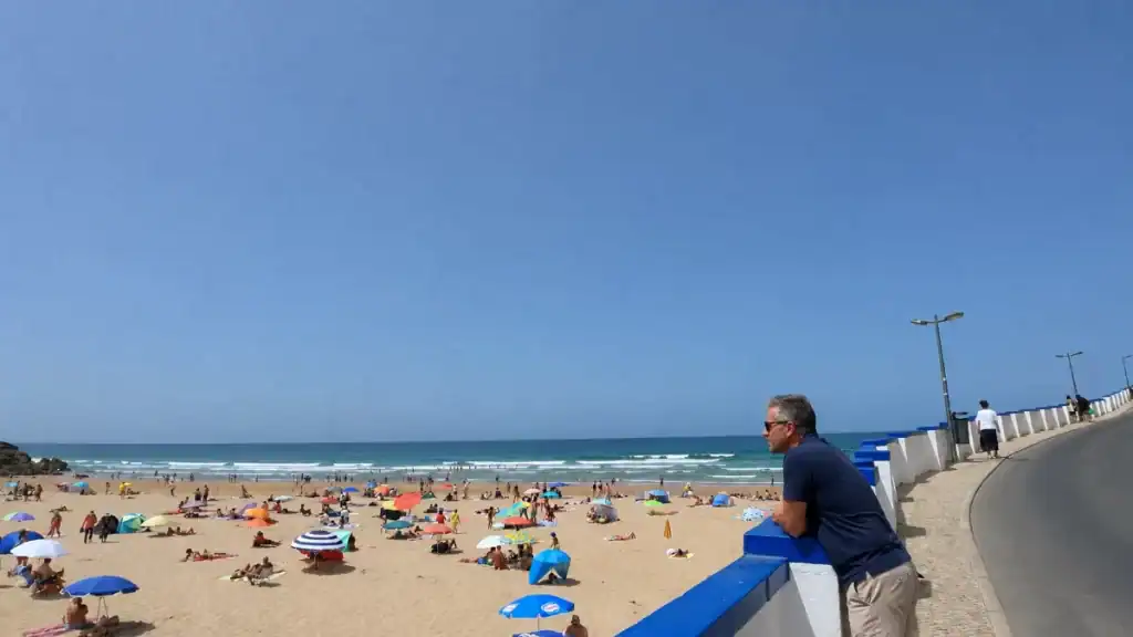 Man leaning on promenade wall overlooking a busy summer beach at Praia das Maçãs