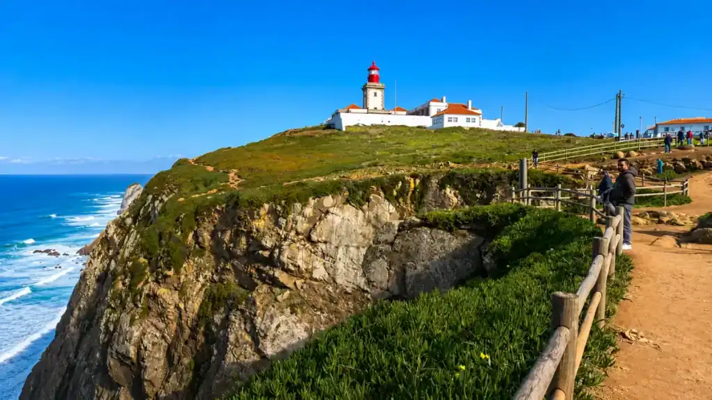 Red-topped lighthouse stands atop the grassy headland of Cabo da Roca above sheer cliffs.