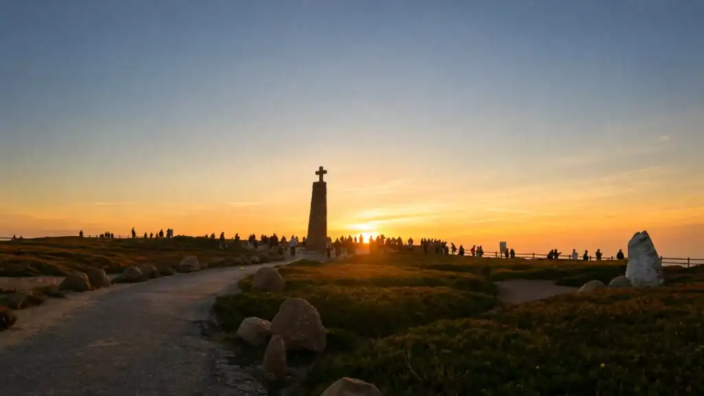 Stone cross monument silhouetted against the setting sun as crowds gather at the cliff edge.