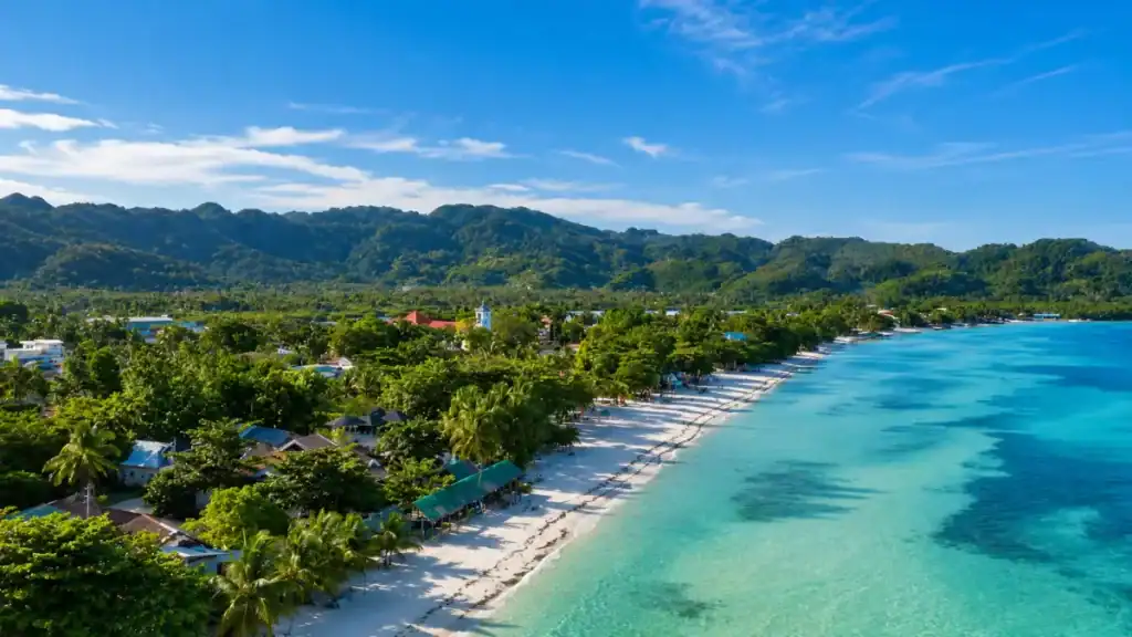 Aerial view shows a long white shoreline meeting turquoise shallows below forested hills at Alona Beach.
