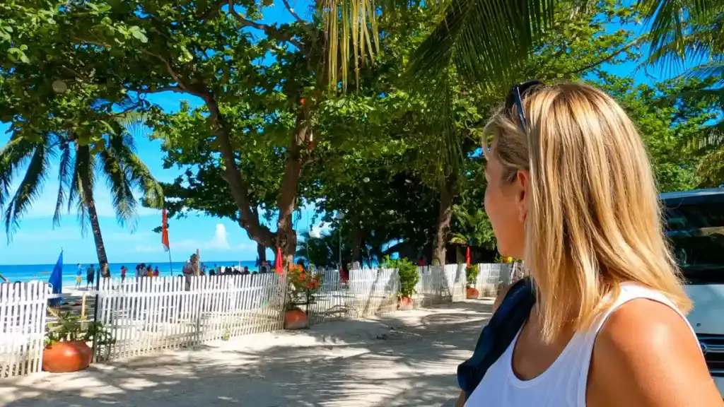 A visitor pauses beneath palm shade beside a white picket fence overlooking Alona Beach.