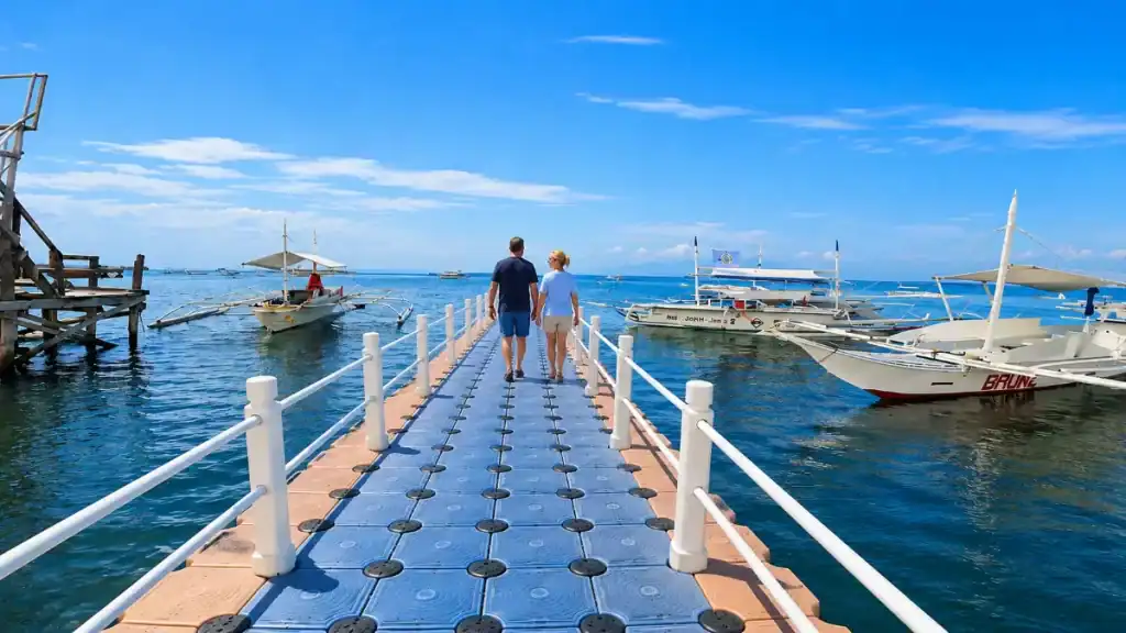 A couple walks a floating pontoon jetty lined with outrigger boats at Panglao port.