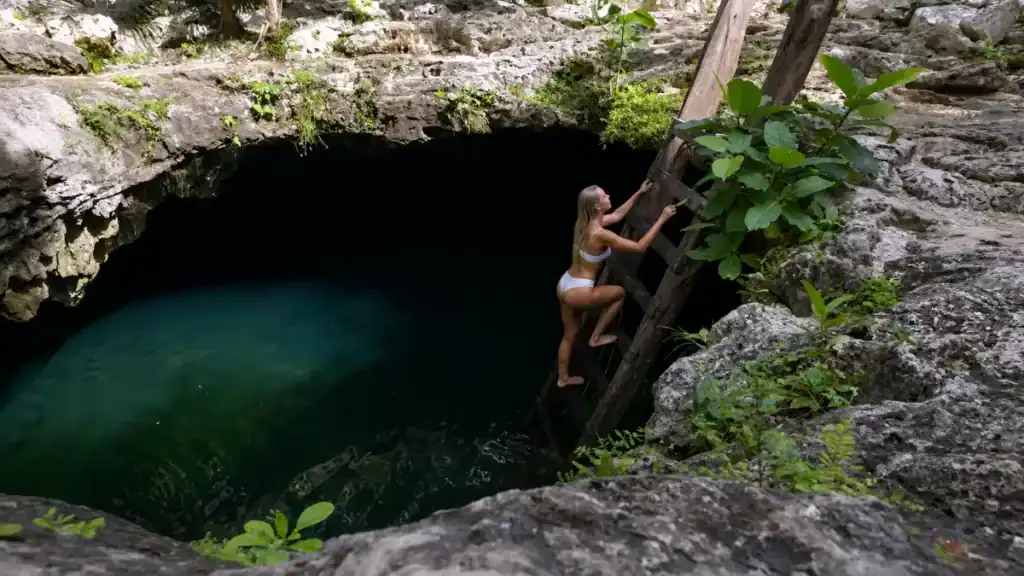 A woman climbs a wooden ladder above Cenote Calavera's dark, cavernous pool.