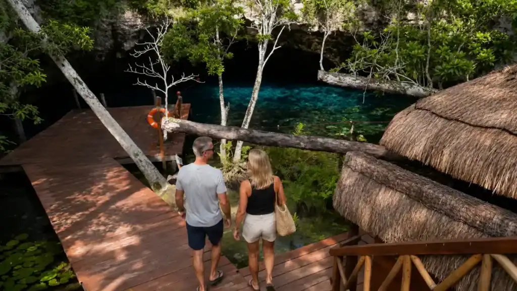 Couple walks a wooden boardwalk toward Gran Cenote's dark blue waters, a free stop worth weighing when deciding is Tulum too expensive.